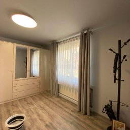 Firma Ogólno-Budowlana Szymon Basisty - Jasne wnętrze pokoju po remoncie: panele podłogowe imitujące drewno, beżowa szafa z lustrami, okno z firanką i zasłonami, wiadro z wodą i mopem, wieszak na ubrania z parasolem i ramką ze zdjęciem.