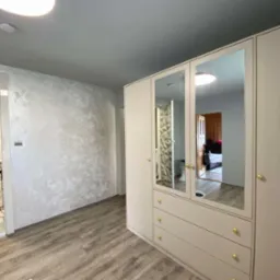 Ściana z dekoracyjną, jasną gładzią w pokoju z drewnopodobną podłogą i szafą z lustrami odbijającymi wnętrze łazienki i sypialni.