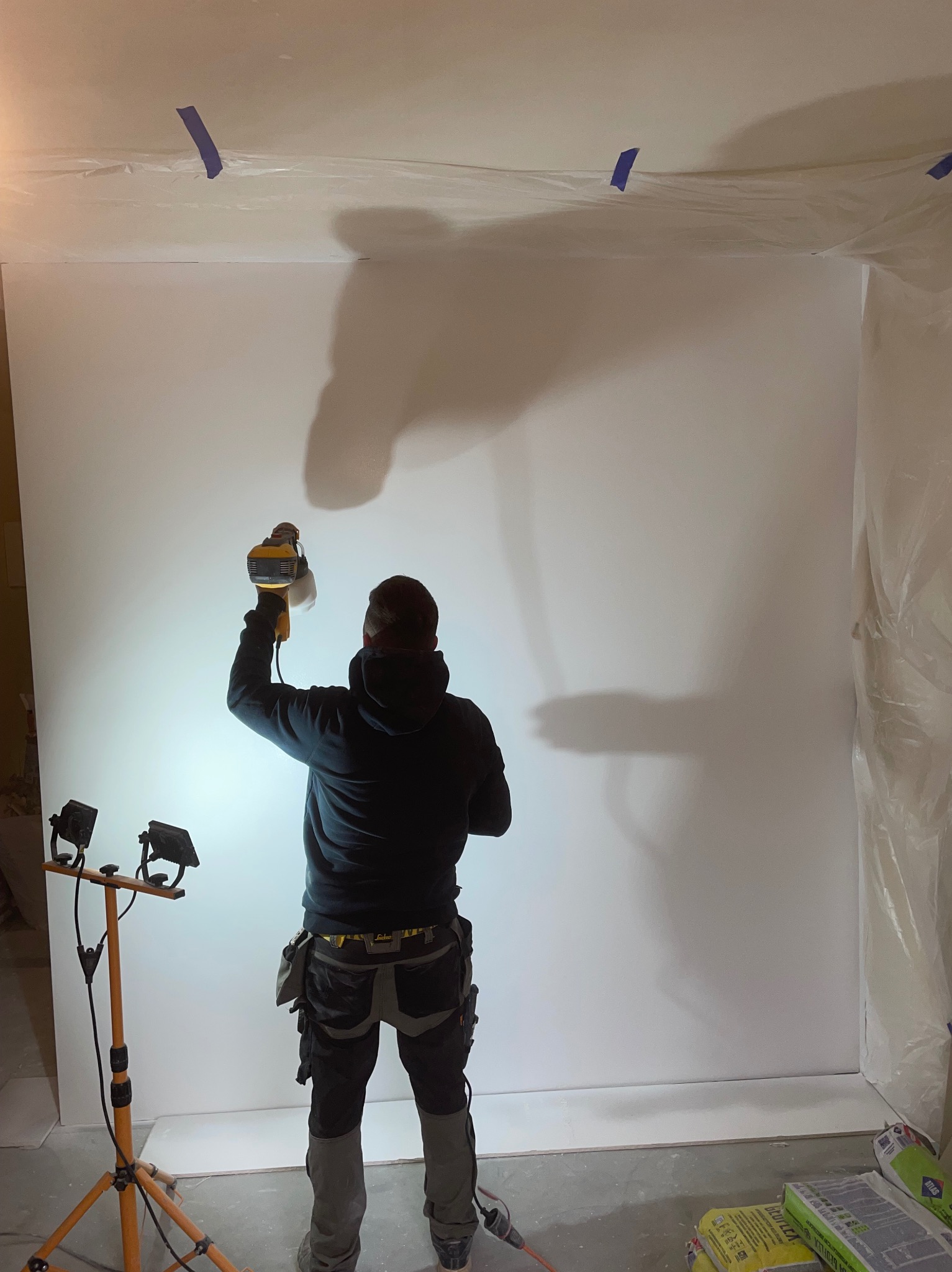 Pracownik w ciemnej bluzie i spodniach roboczych natryskuje białą farbę na ścianę za pomocą elektrycznego pistoletu natryskowego; widoczne taśmy malarskie na suficie i worki zaprawy na podłodze.