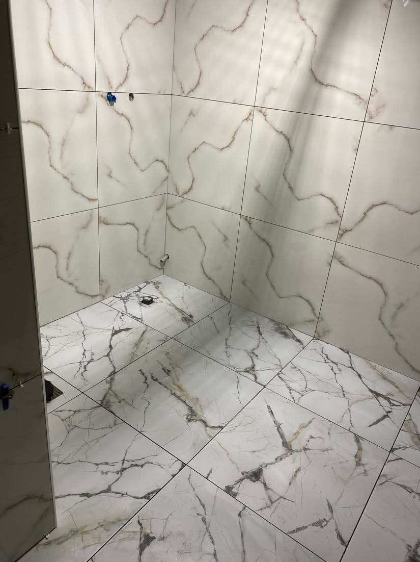 Wykończona kabina prysznicowa z białymi płytkami imitującymi marmur na ścianach i podłodze, widoczne niedokończone przyłącza wodne.