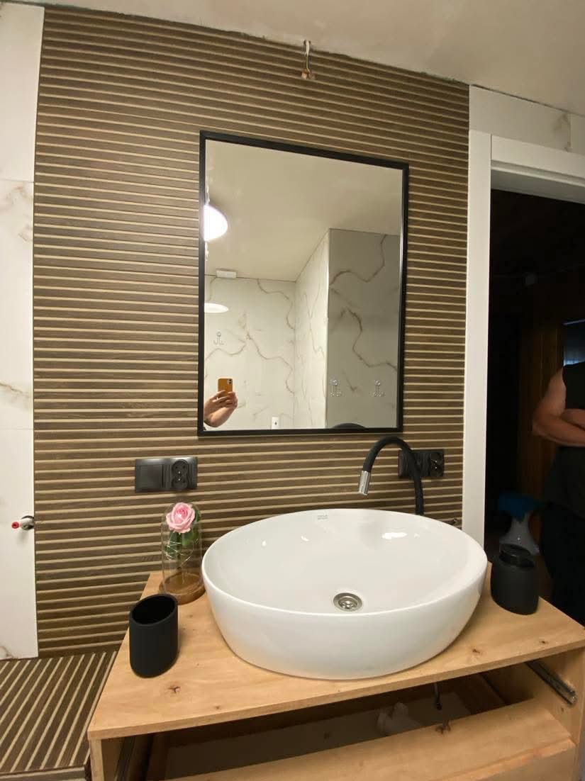 Nowoczesna umywalka nablatowa o okrągłym kształcie na drewnianym blacie, z czarną baterią i dekoracyjną różą pod kloszem, na tle ściany z brązowych paneli dekoracyjnych i lustra z czarną ramą,...