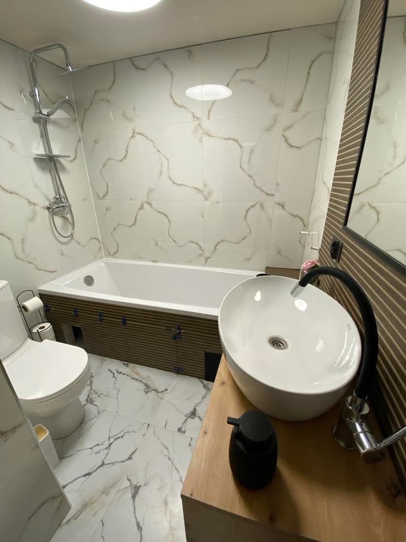 Nowoczesna łazienka z wanną obudowaną drewnianą okładziną, umywalką nablatową na drewnianym blacie i marmurowymi płytkami na ścianach i podłodze.