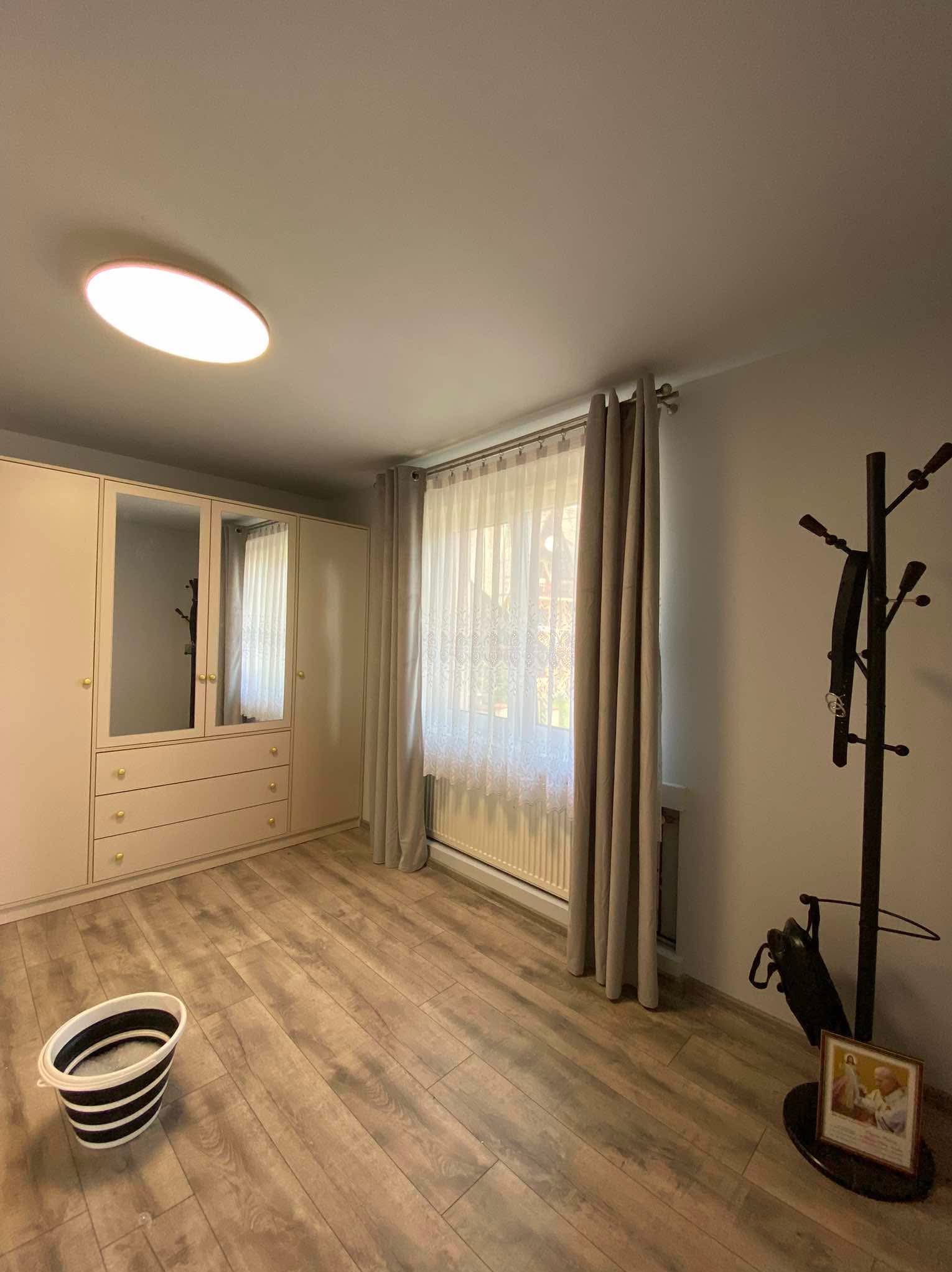 Jasne wnętrze pokoju po remoncie: panele podłogowe imitujące drewno, beżowa szafa z lustrami, okno z firanką i zasłonami, wiadro z wodą i mopem, wieszak na ubrania z parasolem i ramką ze zdjęciem.