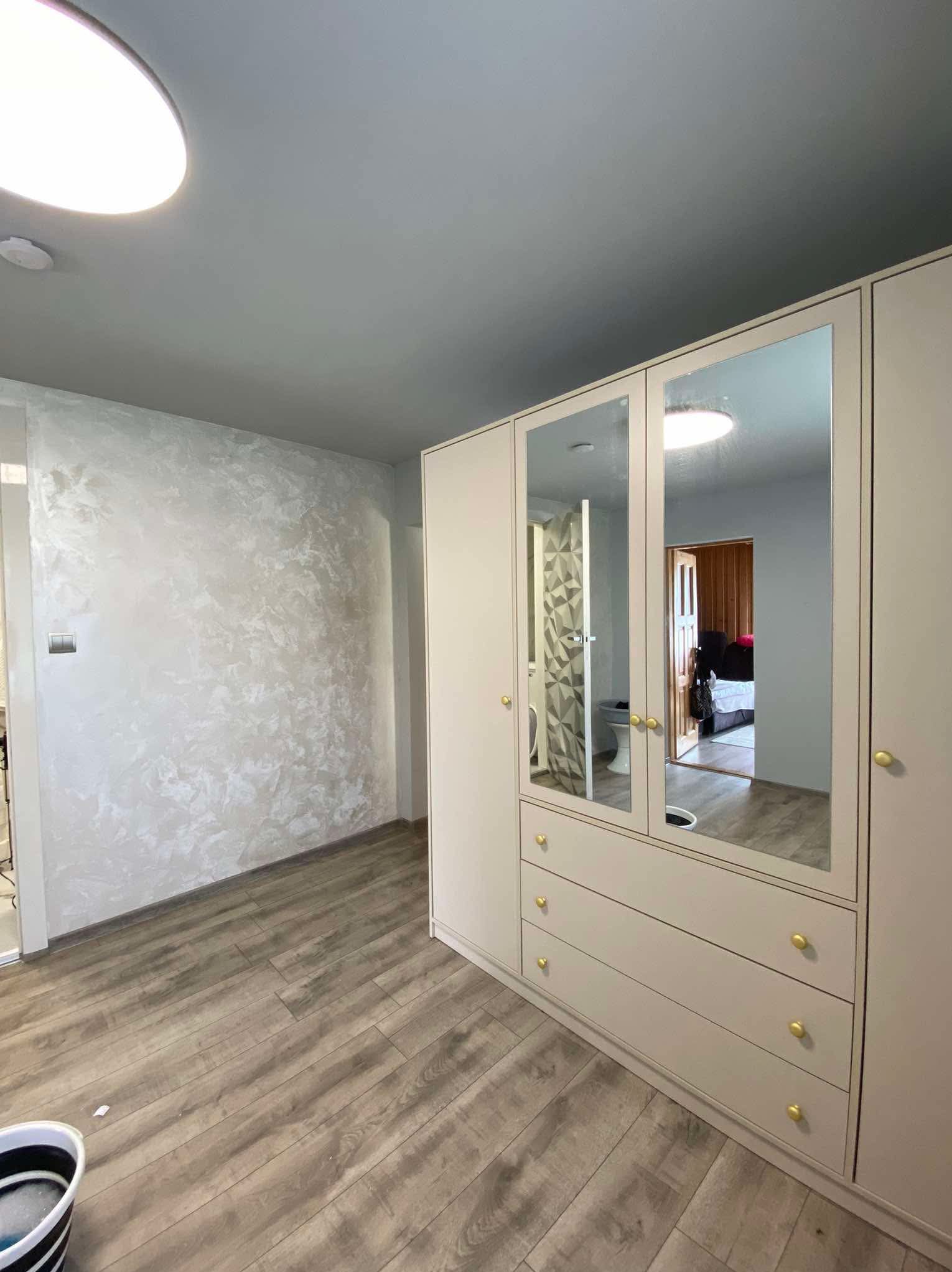 Ściana z dekoracyjną, jasną gładzią w pokoju z drewnopodobną podłogą i szafą z lustrami odbijającymi wnętrze łazienki i sypialni.