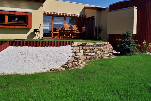 Ogród przydomowy z tarasem, kamiennym murkiem oporowym i rabatą wysypaną białym grysem, widok na budynek z drewnianymi elementami.