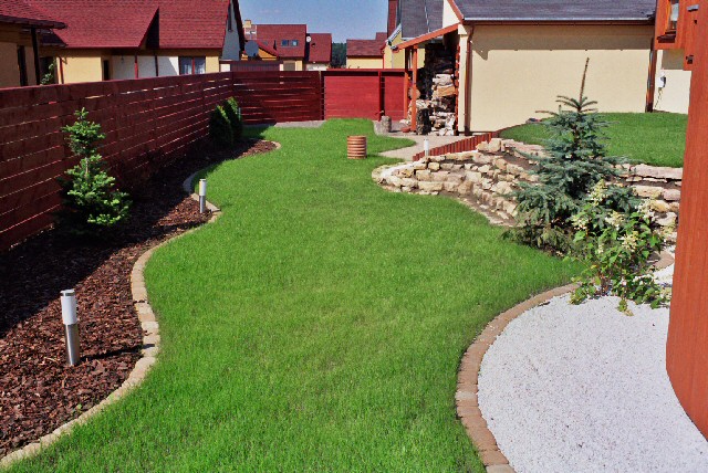 Zadbany ogród z równo przystrzyżonym trawnikiem, obrzeżami z kostki, korą, białym grysem, niską kaskadą z kamieni i młodymi drzewkami iglastymi. Widoczny system nawadniania w postaci cylindrycznego...