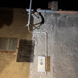 Emil Prochownik - Biała skrzynka elektryczna z żółtym symbolem ostrzegawczym na szorstkiej ścianie budynku, z widocznymi rurami instalacyjnymi i elementami konstrukcyjnymi.