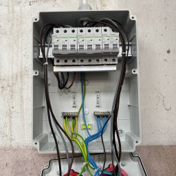 Emil Prochownik - Otwarta skrzynka elektryczna z bezpiecznikami Noark, widoczne przewody w kolorach: niebieskim, żółto-zielonym, brązowym i czarnym. Poziomica wewnątrz.
