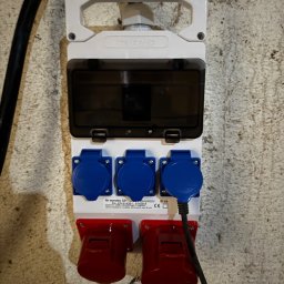 Emil Prochownik - Rozdzielnica elektryczna natynkowa z gniazdami siłowymi (czerwone i niebieskie) zamontowana na szarej, chropowatej ścianie. Widoczny kabel zasilający.
