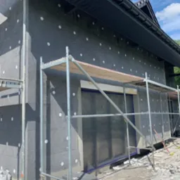 Dom w trakcie ocieplania styropianem, widoczne rusztowania, zabezpieczone okna folią, porozrzucane fragmenty styropianu na ziemi.