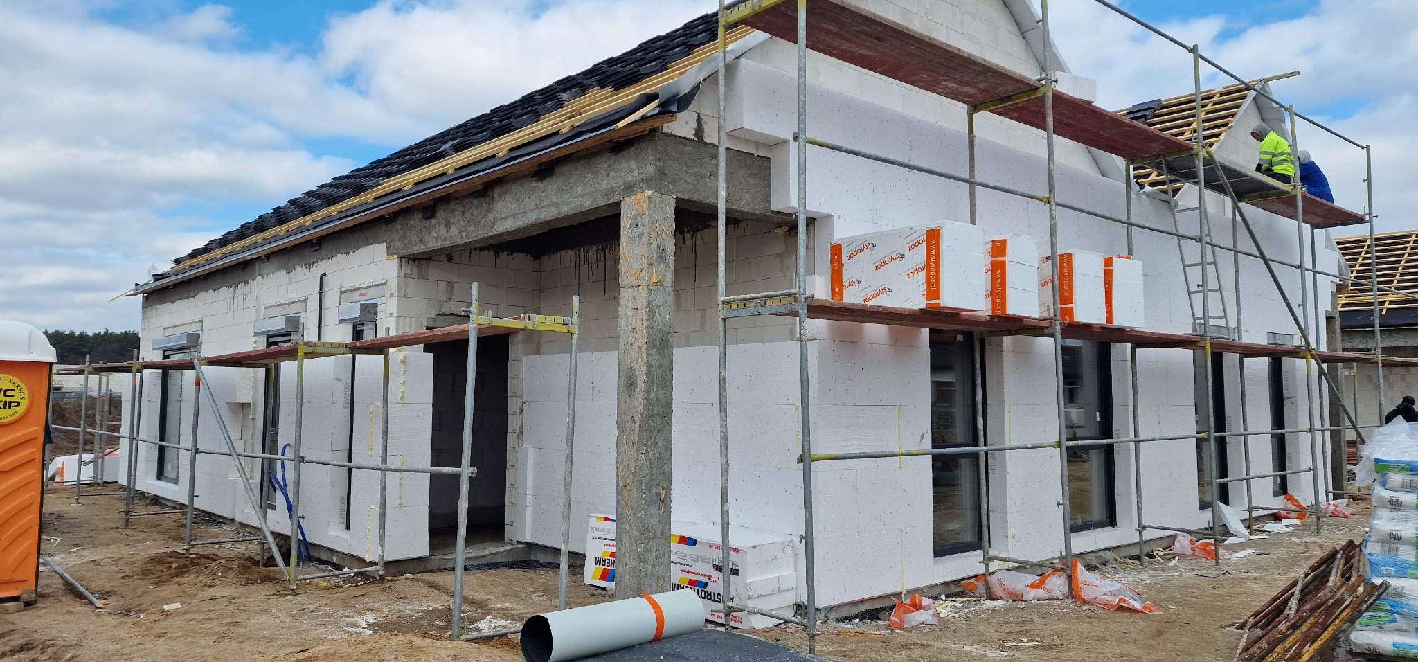 Budowa domu jednorodzinnego w trakcie ocieplania ścian styropianem, widoczne rusztowania, niezabezpieczone okna, porozrzucane materiały budowlane i pracownicy na dachu