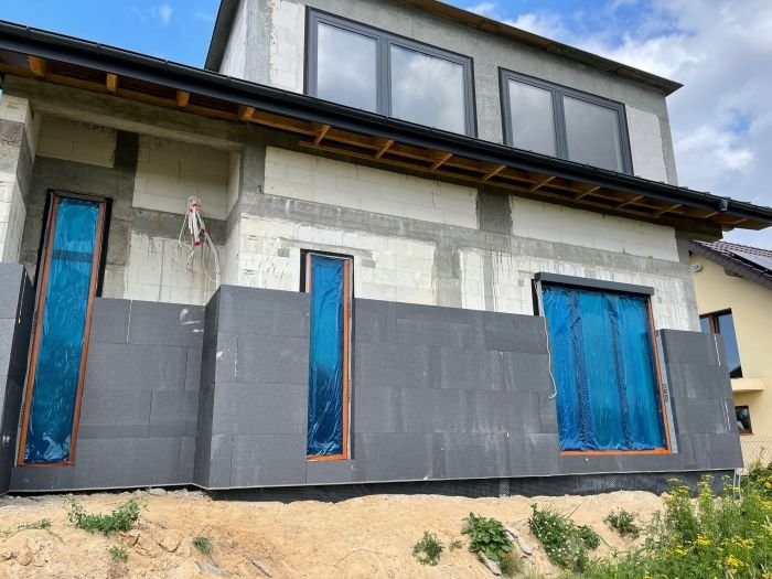 Dom w trakcie prac izolacyjnych: szare płyty styropianowe na ścianie, okna zabezpieczone folią, widoczne fragmenty surowego muru i instalacji elektrycznej.