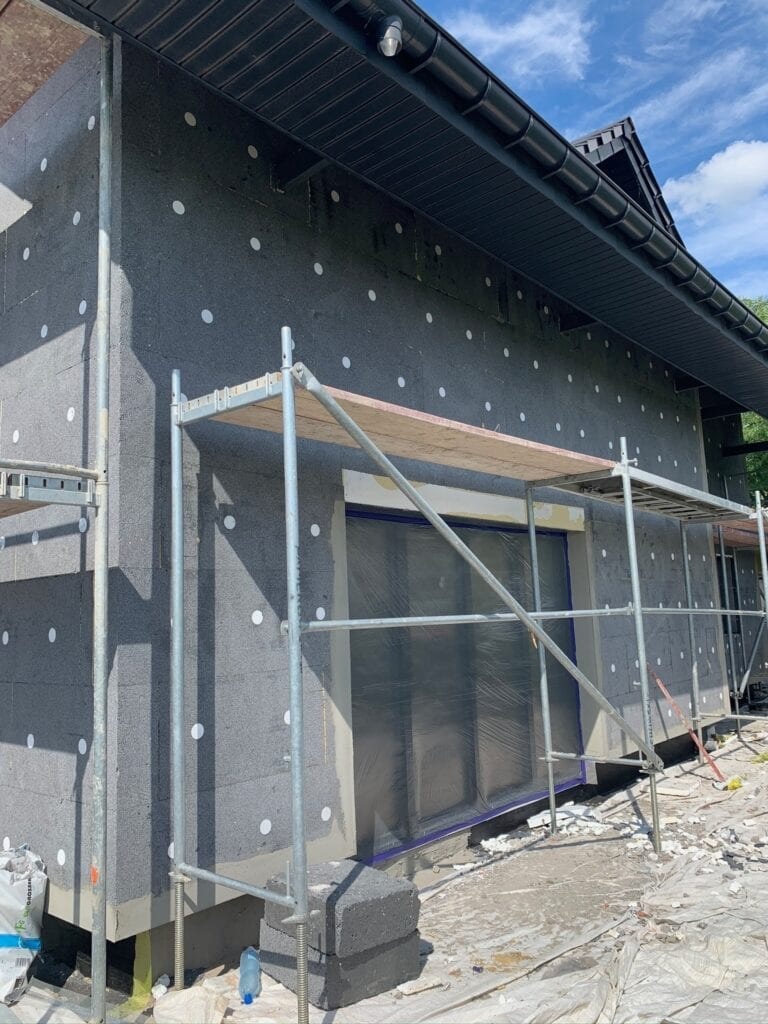 Dom w trakcie ocieplania styropianem, widoczne rusztowania, zabezpieczone okna folią, porozrzucane fragmenty styropianu na ziemi.