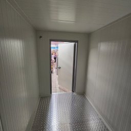 Arbilem Daria Piechota - Wnętrze chłodni z otwartymi drzwiami, widoczna metalowa podłoga z wzorem i białe panele ścienne. W tle osoba w sklepie.