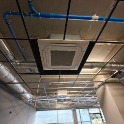 Arbilem Daria Piechota - Widok z dołu na zamontowaną kasetę sufitową klimatyzacji w biurze, z widocznymi elementami instalacji wentylacyjnych i rurami. Oświetlenie dzienne przez okna.