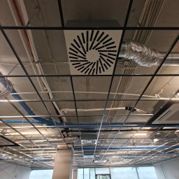 Arbilem Daria Piechota - Sufit biurowy z widocznymi instalacjami: wentylacyjnymi, elektrycznymi i klimatyzacyjnymi. Widoczne kratki wentylacyjne o radialnym wzorze i rury wentylacyjne.