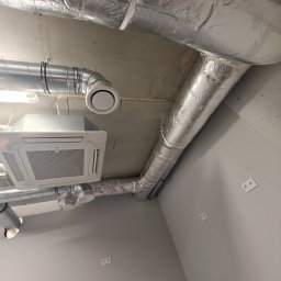 Arbilem Daria Piechota - Srebrne rury wentylacyjne i biała kaseta klimatyzacyjna zamontowane pod betonowym sufitem w pomieszczeniu o szarych ścianach z gniazdkami elektrycznymi.