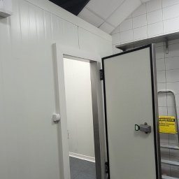 Arbilem Daria Piechota - Wnętrze chłodni z otwartymi drzwiami, widoczna drabina i wózek transportowy. Białe płytki na ścianach i sufit, metalowa półka. Oświetlenie sufitowe.