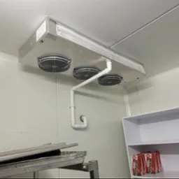 Zamontowana sufitowa chłodnica przemysłowa z trzema wentylatorami w chłodni, z widocznymi rurami odprowadzającymi skropliny i półkami z produktami.