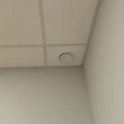 Biały, okrągły nawiewnik wentylacyjny w suficie kasetonowym, widoczny z dołu pod kątem. Szara ściana w tle. Minimalistyczny kadr, neutralne oświetlenie.