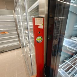 Arbilem Daria Piechota - Czerwona poziomica przyłożona do metalowej ramy szklanej chłodziarki sklepowej, sprawdzanie wypoziomowania urządzenia. Widoczne puste półki chłodziarki.