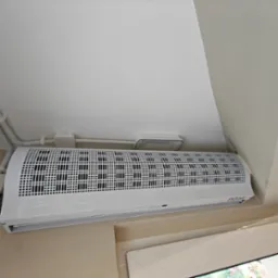 Biała kurtyna powietrzna NOXA zamontowana nad oknem w Częstochowie. Widoczne rury instalacyjne i lampa sufitowa. Białe ściany i okno.