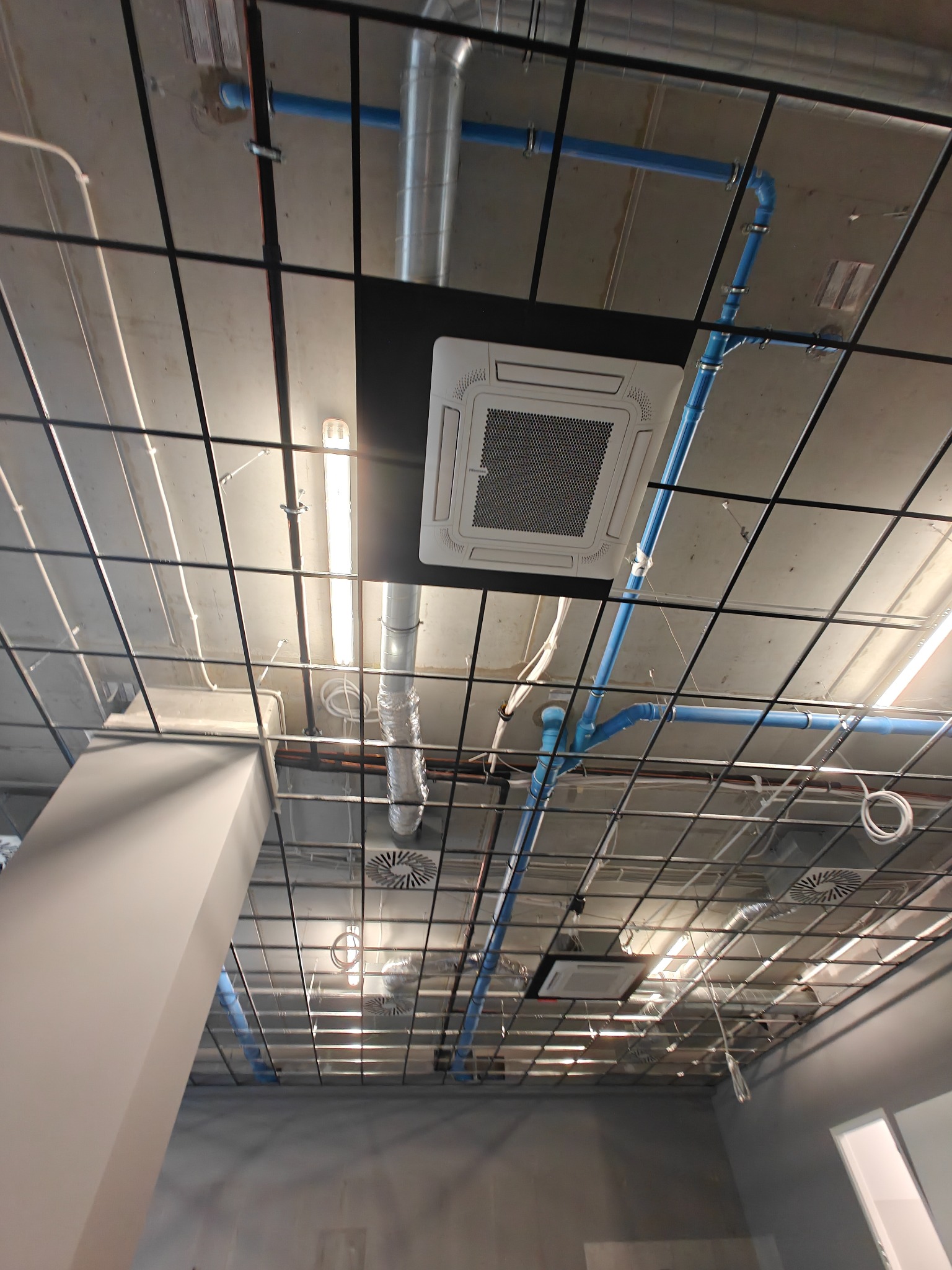 Sufit biurowy z widocznymi instalacjami: klimatyzacja kasetonowa, rury wentylacyjne, przewody elektryczne, oświetlenie liniowe. Metalowa konstrukcja podtrzymująca.