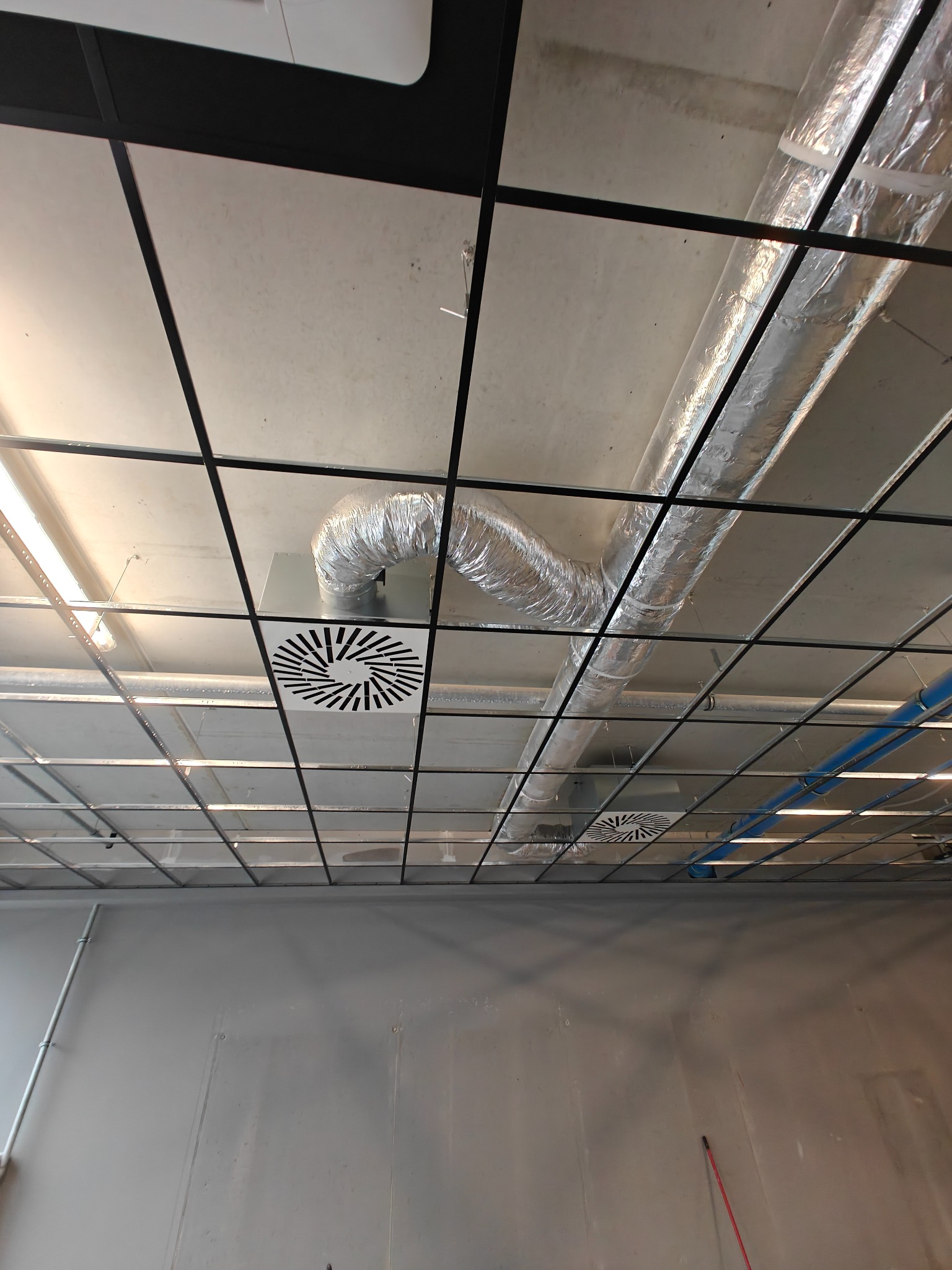Srebrny kanał wentylacyjny i okrągły dyfuzor w rastrowym suficie biurowym z widocznymi instalacjami. Widok z dołu.