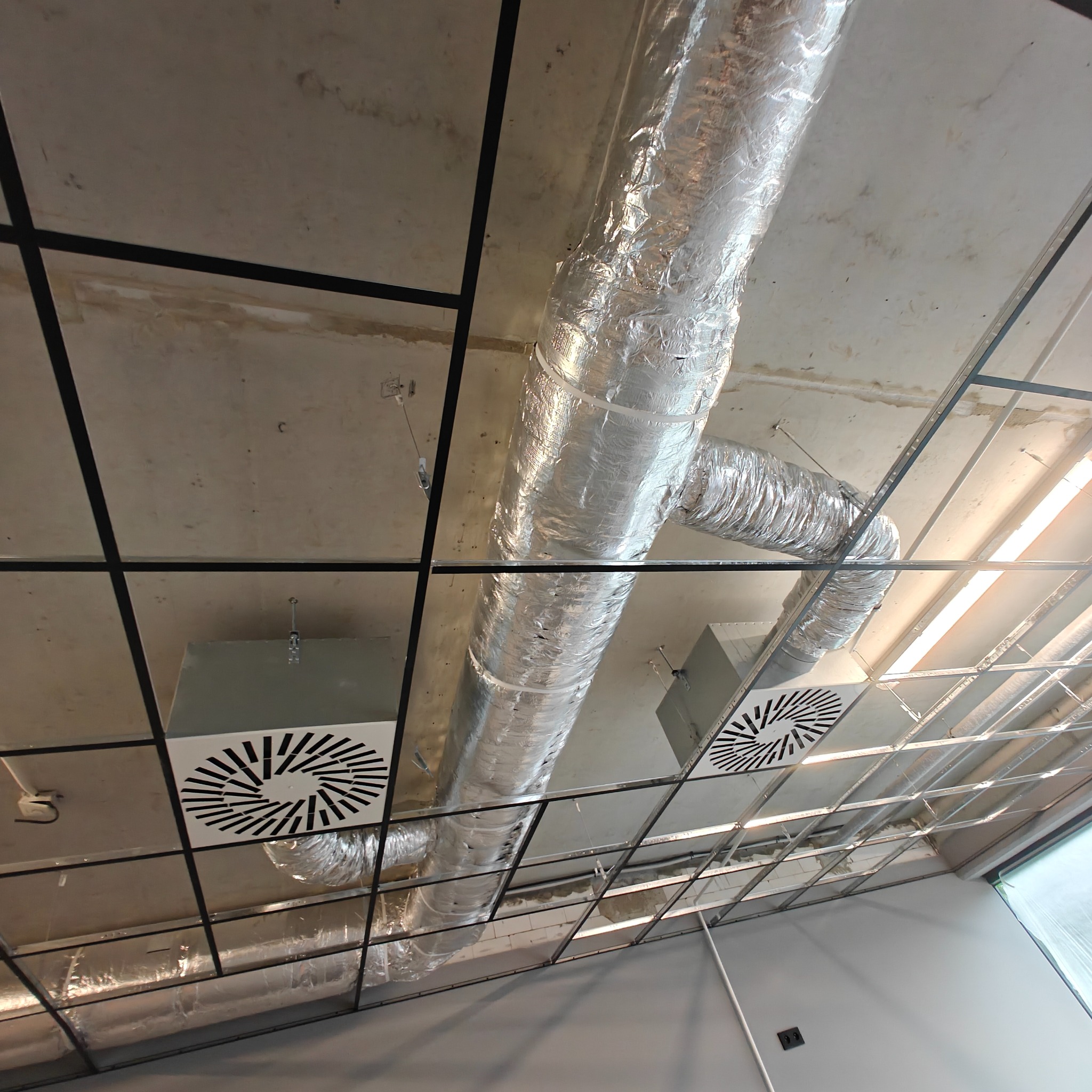 Srebrny, izolowany kanał wentylacyjny z kratkami w rastrowym suficie podwieszanym, widok z dołu. Elementy instalacji wentylacyjnej w biurze.