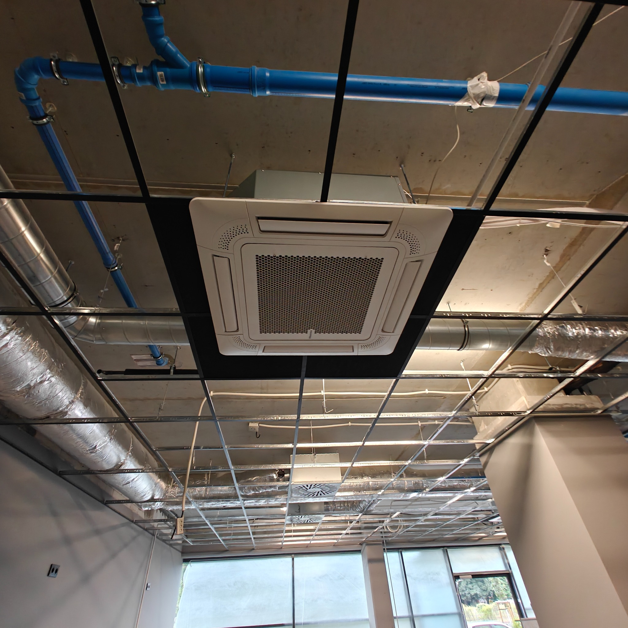Widok z dołu na zamontowaną kasetę sufitową klimatyzacji w biurze, z widocznymi elementami instalacji wentylacyjnych i rurami. Oświetlenie dzienne przez okna.