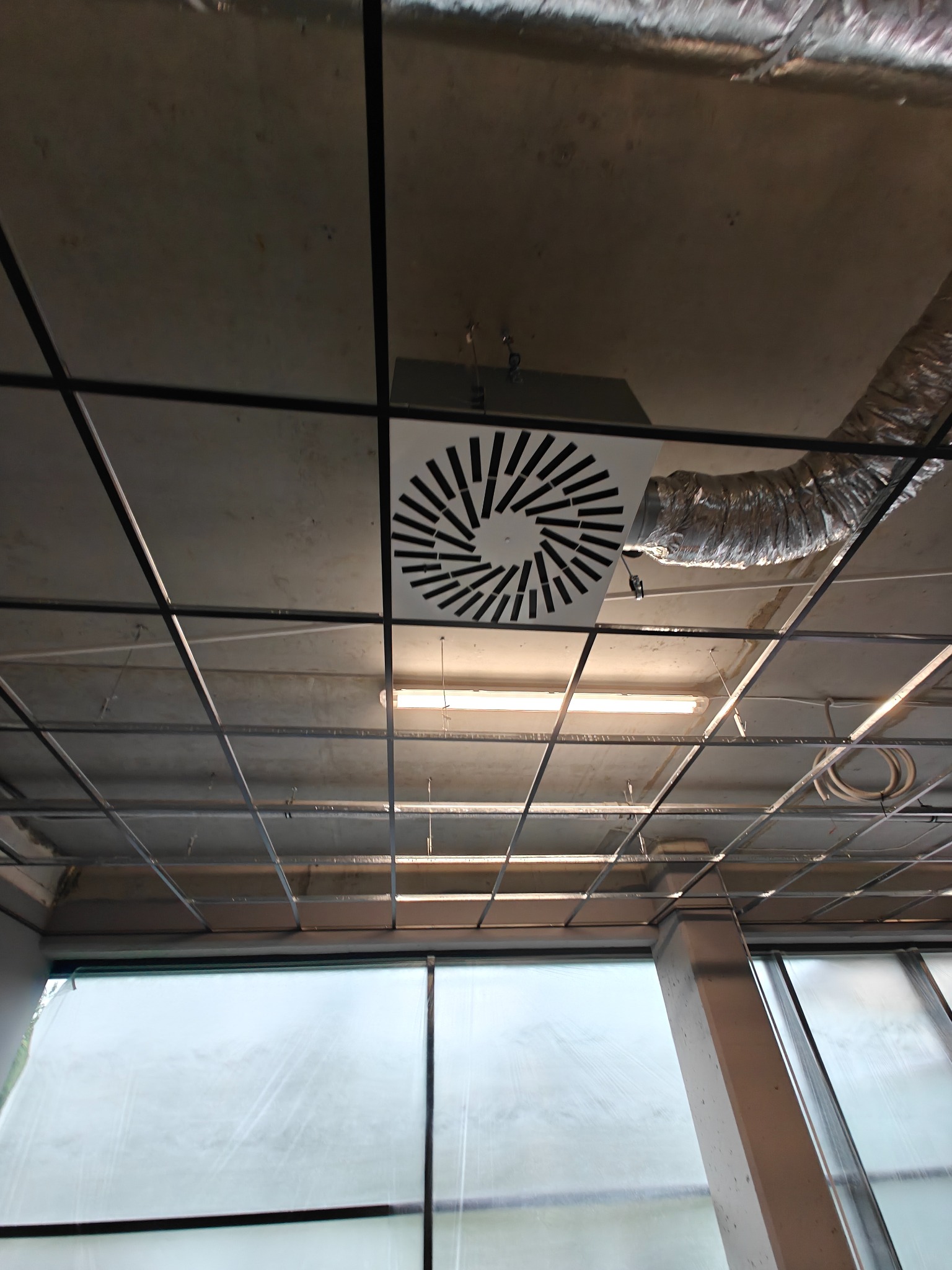 Widok z dołu na zamontowany nawiewnik sufitowy z okrągłym wzorem, podwieszony sufit rastrowy i fragment instalacji wentylacyjnej w pomieszczeniu biurowym.