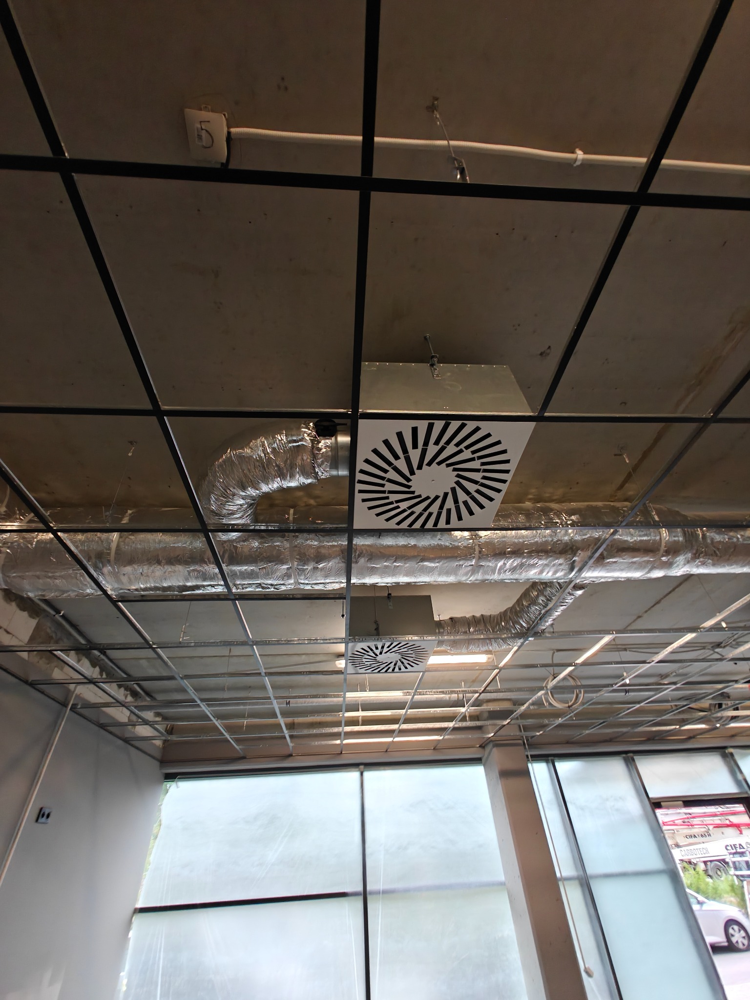 Widok z dołu na zamontowane, białe nawiewniki wentylacyjne z dekoracyjnym wzorem na suficie w budynku komercyjnym z widocznymi rurami wentylacyjnymi i oknami.