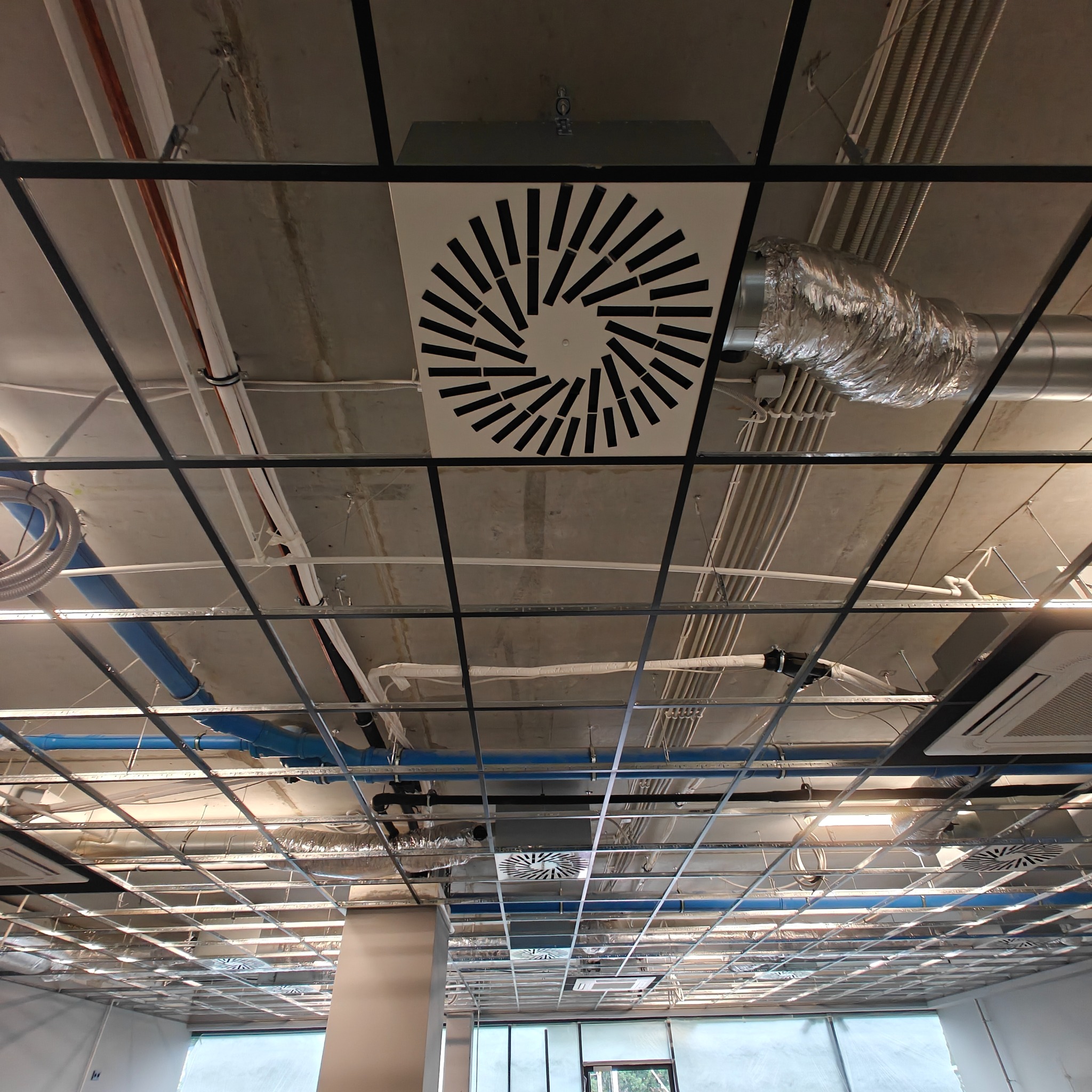 Sufit biurowy z widocznymi instalacjami: wentylacyjnymi, elektrycznymi i klimatyzacyjnymi. Widoczne kratki wentylacyjne o radialnym wzorze i rury wentylacyjne.