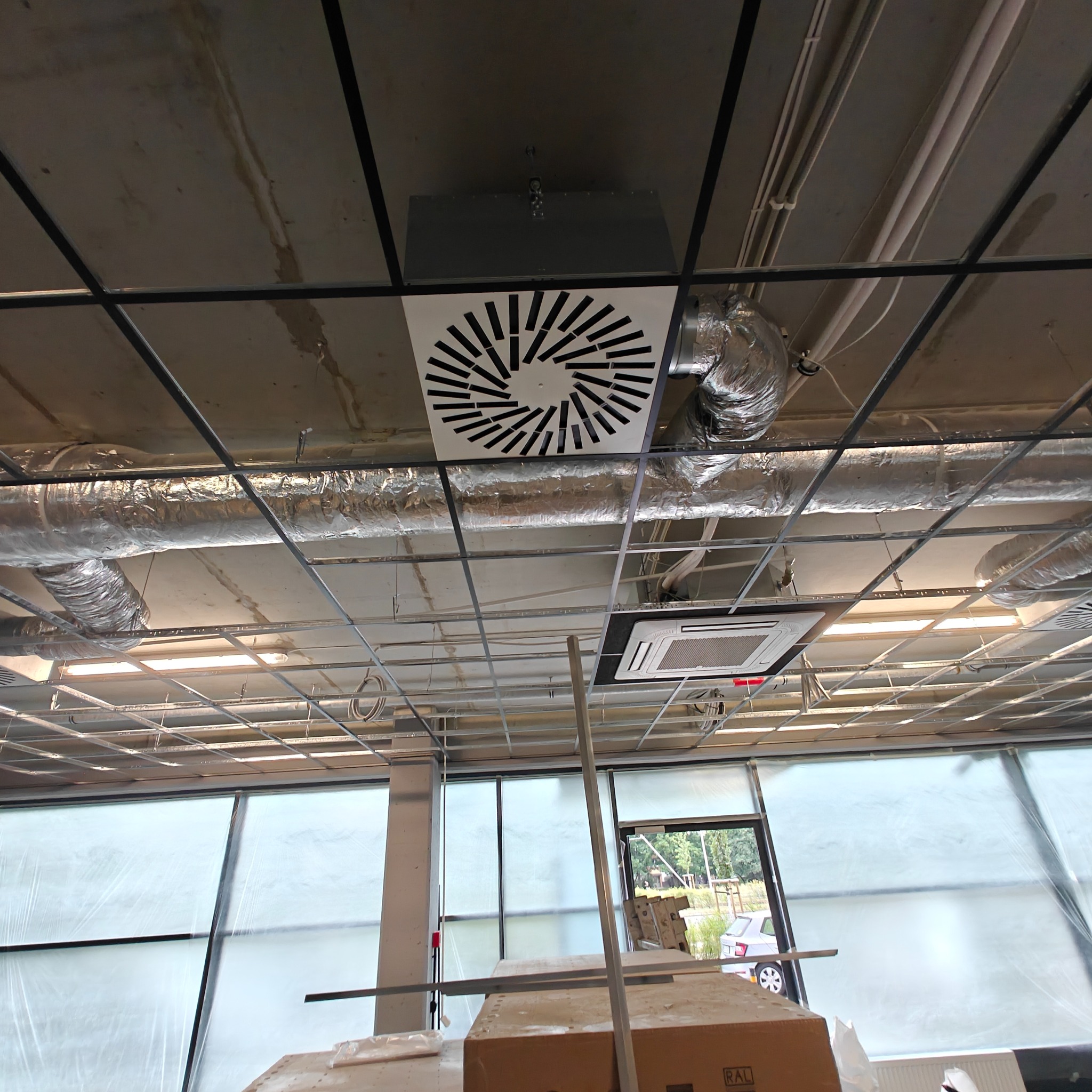 Sufit biura w trakcie montażu wentylacji: widoczny system rur, kratka wentylacyjna z wzorem radialnym i klimatyzator kasetonowy. Okna zasłonięte folią.