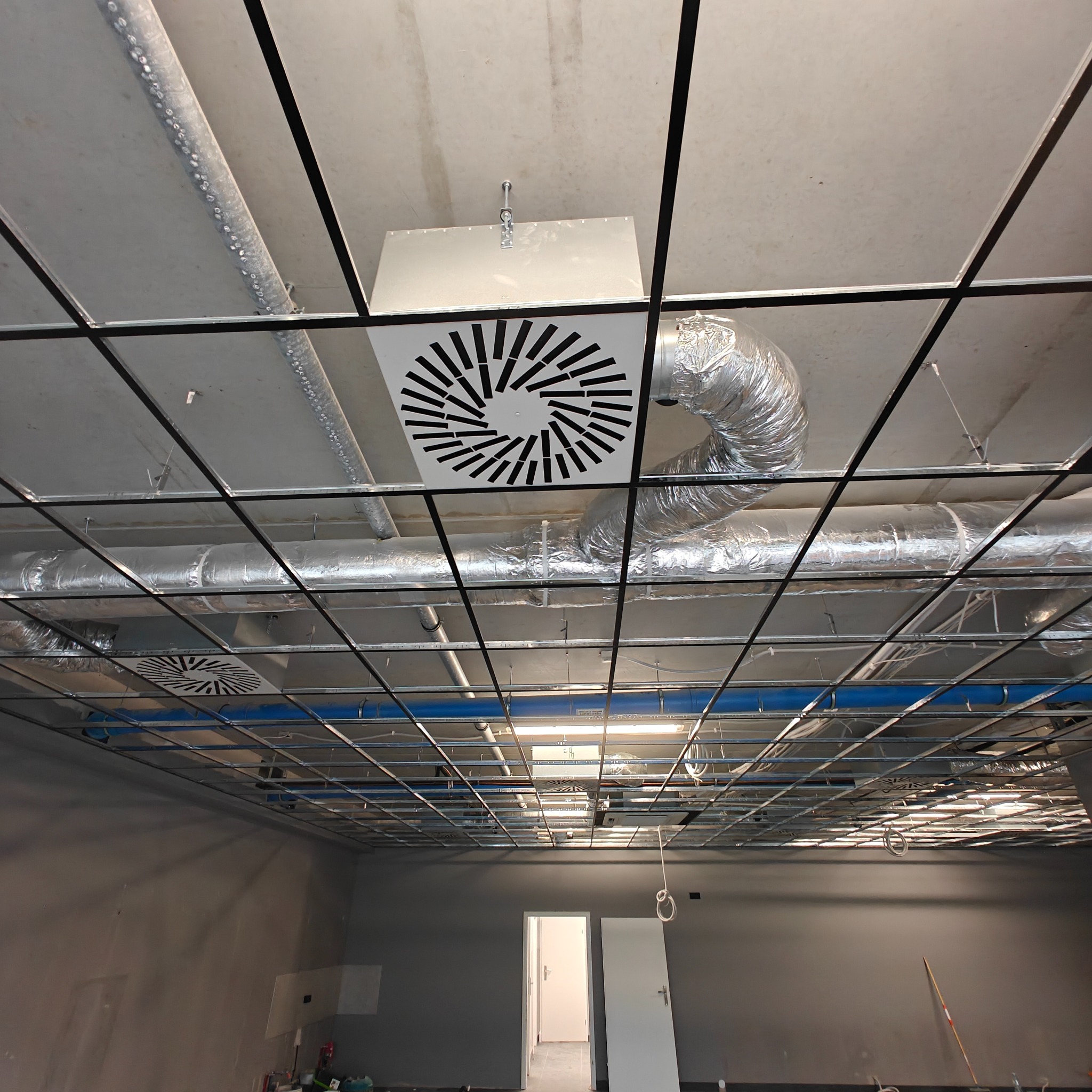 Widok z dołu na sufit z zamontowaną instalacją wentylacyjną z aluminiowymi rurami i kratkami wentylacyjnymi w nowym biurze. Widoczna metalowa konstrukcja sufitu podwieszanego.