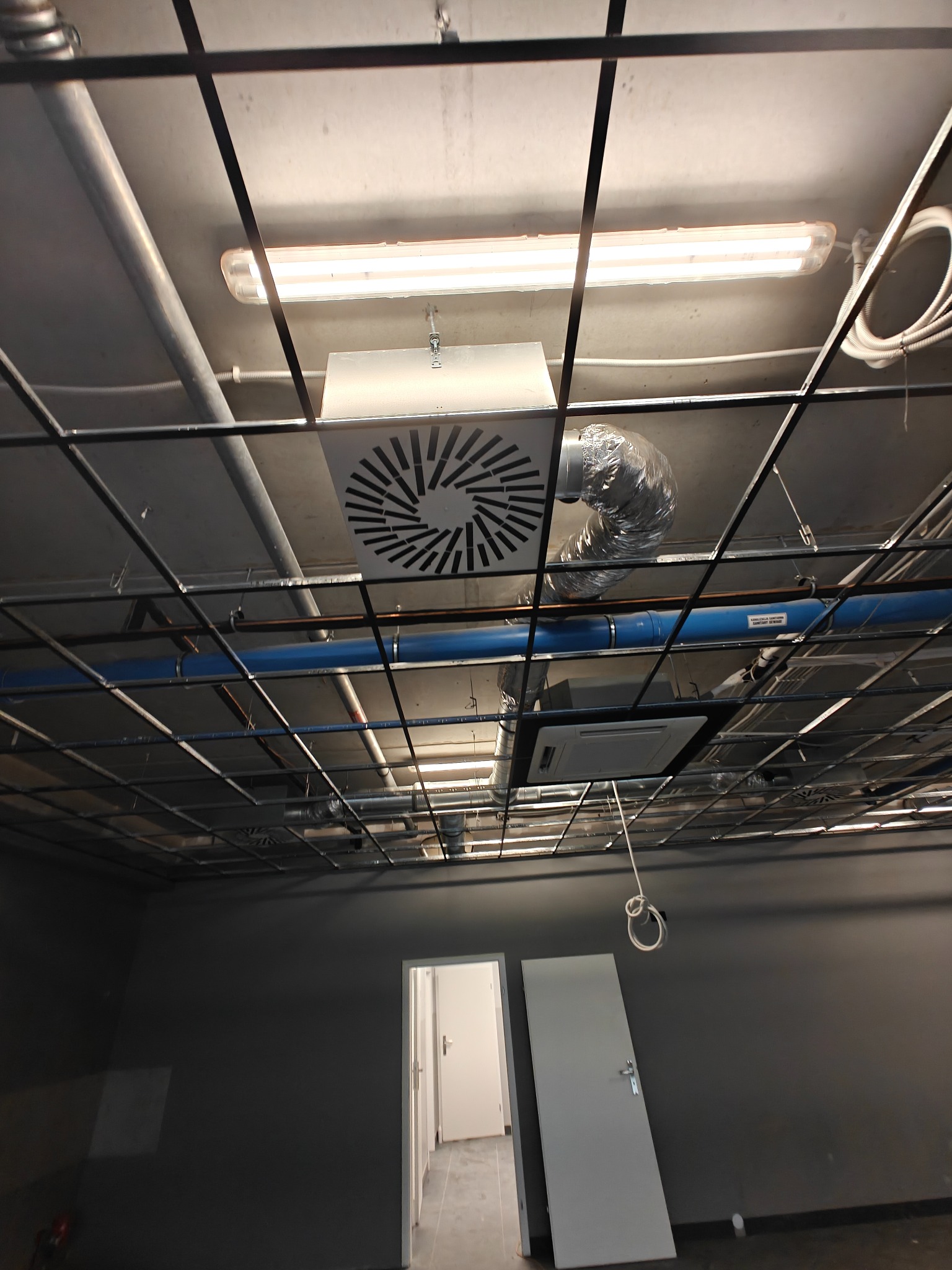 Sufit biura z widoczną instalacją wentylacyjną: kanał wentylacyjny, kratka nawiewna, rury i oświetlenie. Widok z dołu na szarą ścianę i drzwi.