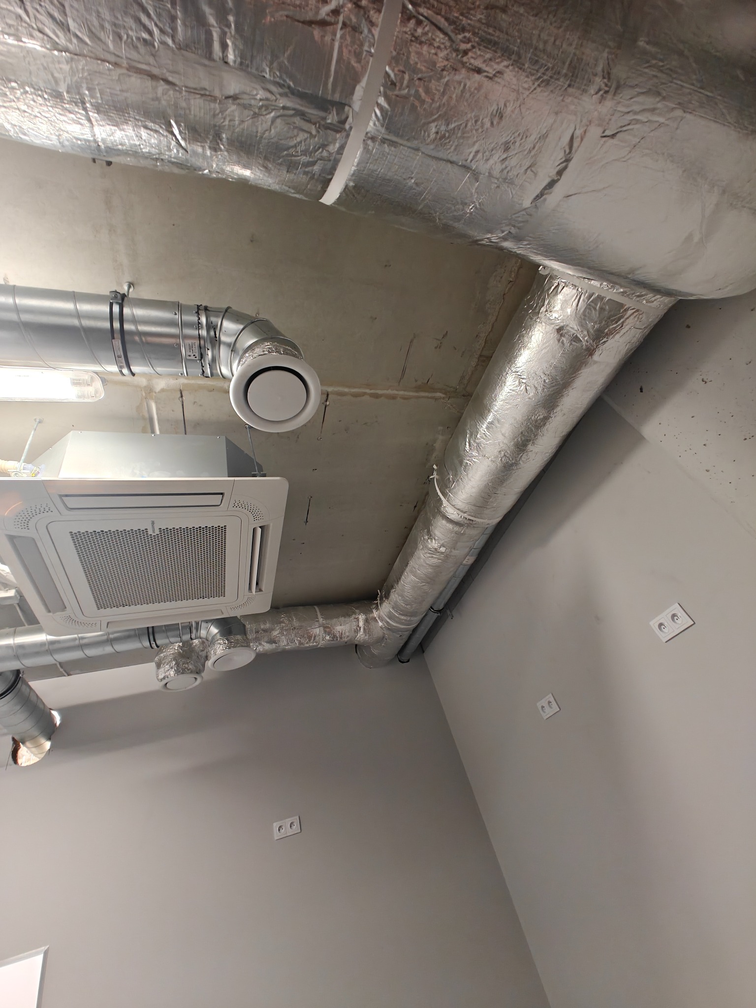 Srebrne rury wentylacyjne i biała kaseta klimatyzacyjna zamontowane pod betonowym sufitem w pomieszczeniu o szarych ścianach z gniazdkami elektrycznymi.