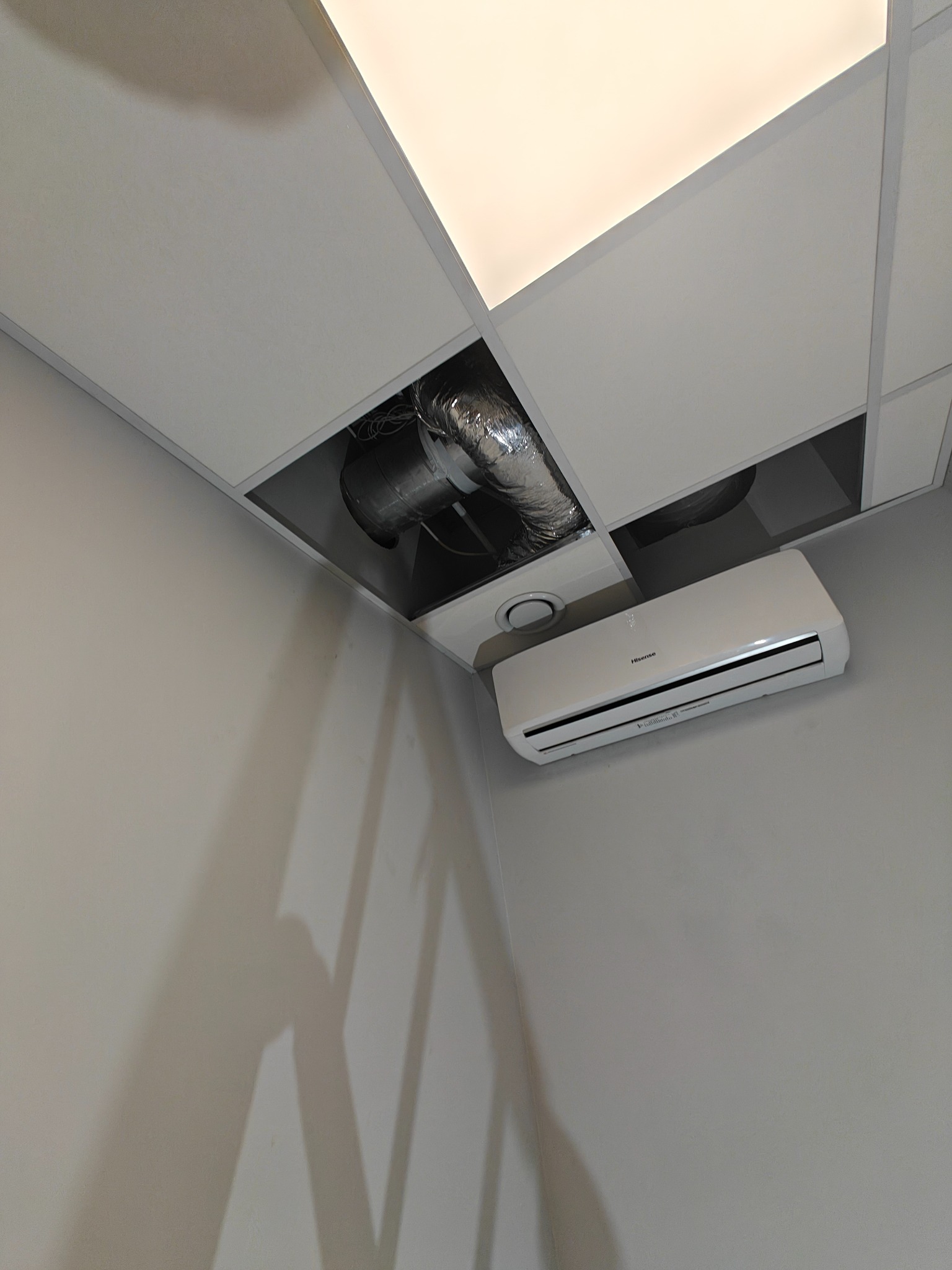 Biały klimatyzator Hisense zamontowany pod sufitem kasetonowym z widocznymi elementami instalacji wentylacyjnej. Szare ściany, ujęcie z dołu.