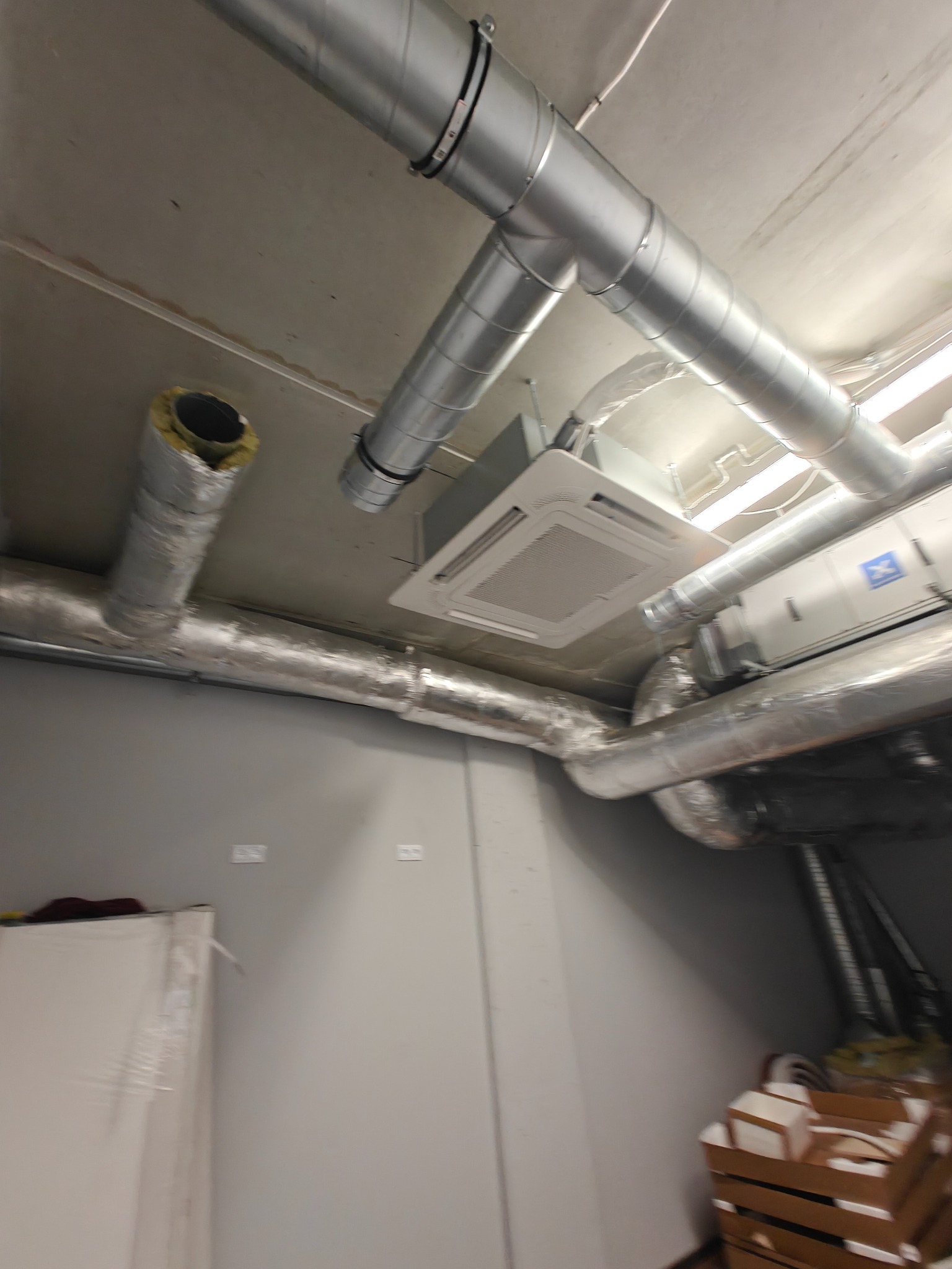 Srebrne rury wentylacyjne i biała klimatyzacja kasetonowa pod sufitem w jasnym pomieszczeniu. Widoczne łączenia i elementy instalacji w przestrzeni komercyjnej.