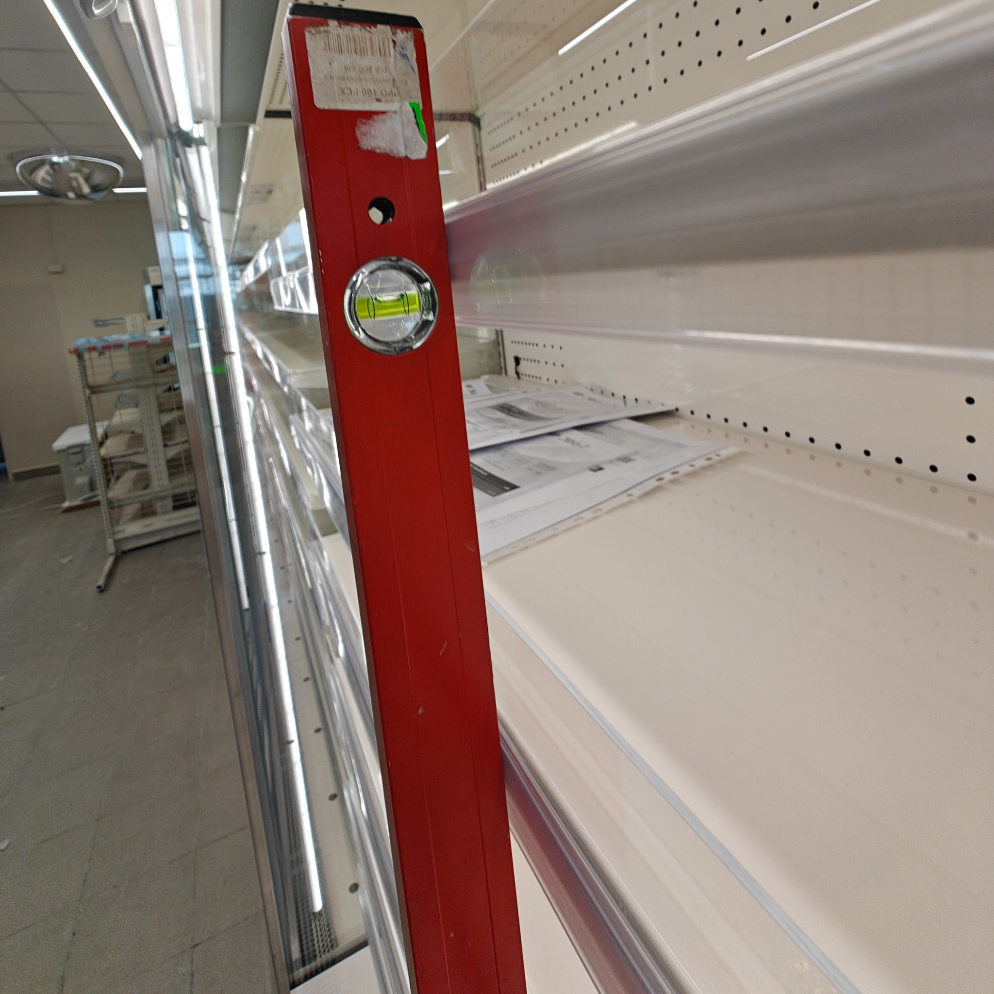 Czerwona poziomica przyłożona do pustej półki sklepowej, sprawdzanie poziomu, widoczne otwory montażowe. W tle widoczny regał z wózkami.