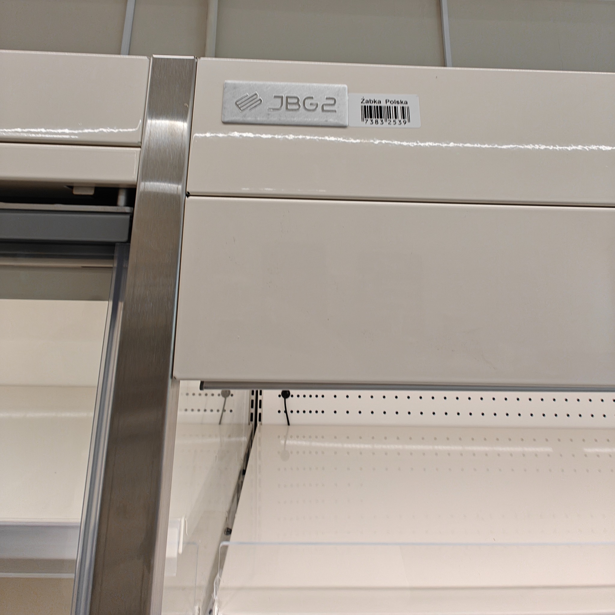 Półka chłodnicza JBG2 w sklepie Żabka Polska, widok z boku, pusta. Oświetlenie górne. Widoczny kod kreskowy i logo producenta.