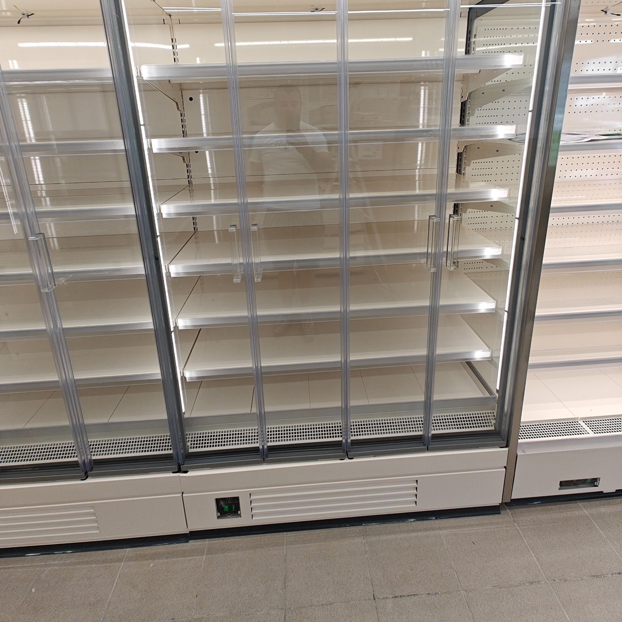 Puste, chłodnicze regały sklepowe z przeszklonymi drzwiami. Widoczne półki i oświetlenie wewnątrz. Podłoga z płytek w neutralnym kolorze.