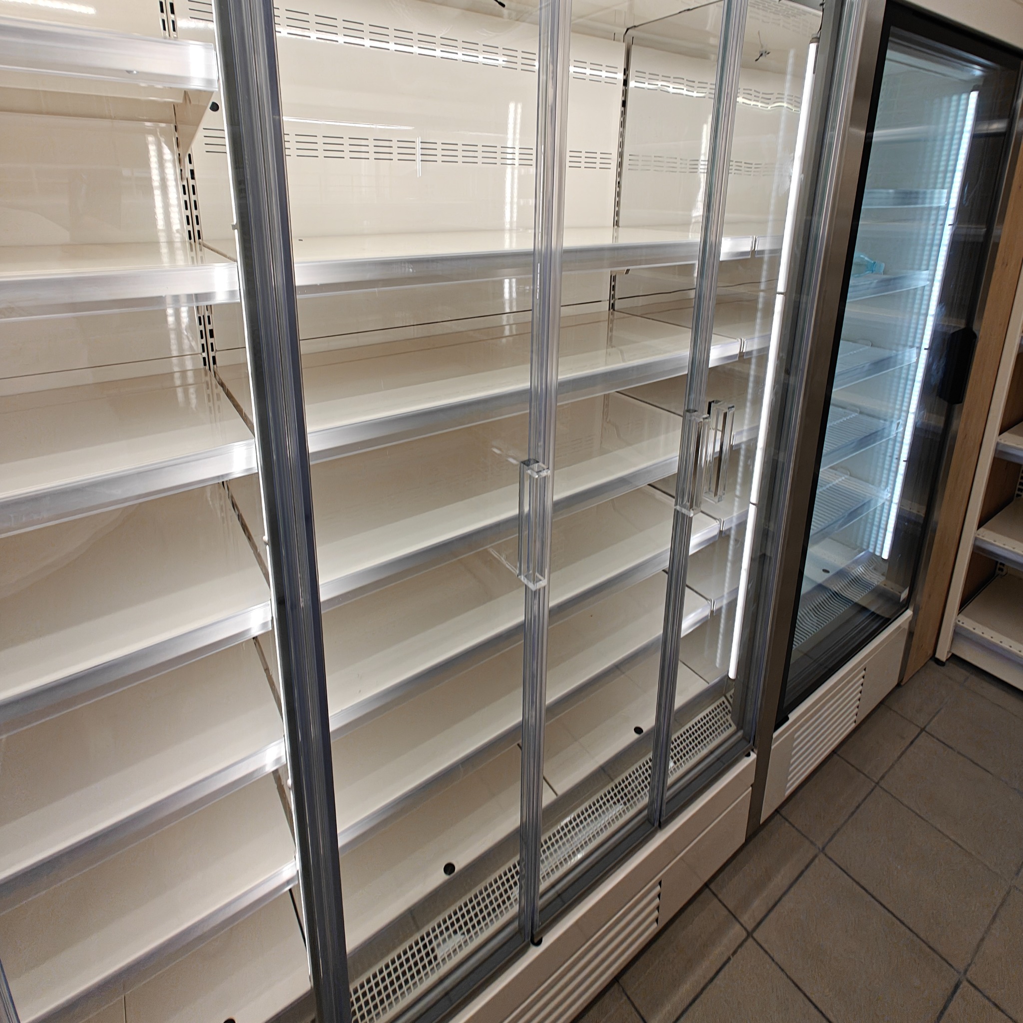 Pusta chłodziarka sklepowa z przesuwnymi szklanymi drzwiami, widoczne półki i oświetlenie wewnętrzne. Widok z boku, ukazujący detale konstrukcyjne.