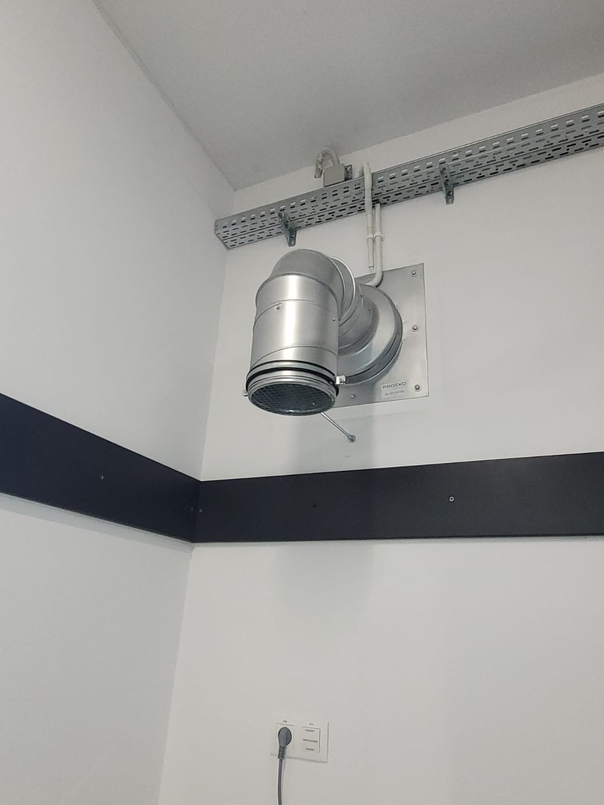 Srebrny anemostat sufitowy z kratką, zamontowany na białej ścianie, z widoczną listwą kablową i czarnym pasem ochronnym. Ujęcie w narożniku pomieszczenia.