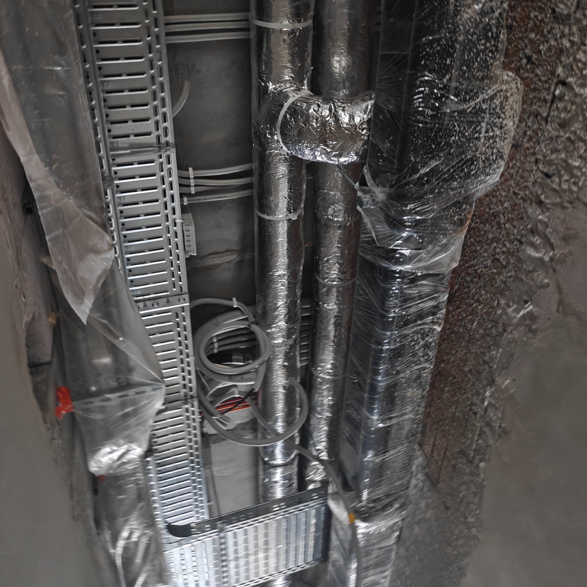 Szare rury wentylacyjne z folią aluminiową i kable w korytkach na suficie budynku w trakcie montażu, widok z dołu, oświetlenie techniczne.