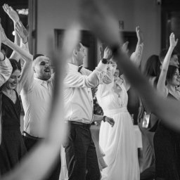 Aleksander Gajda - Czarno-białe zdjęcie grupy roześmianych osób w eleganckich strojach, z uniesionymi rękami, prawdopodobnie podczas tańca na przyjęciu.