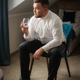 Aleksander Gajda - Mężczyzna w białej koszuli i ciemnych spodniach siedzi na turkusowym krześle, trzymając szklankę wody i spoglądając w dal, w tle widoczne łóżko z ciemnymi poduszkami.