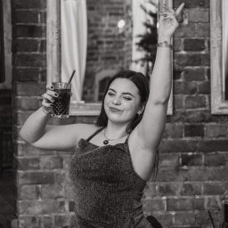 Aleksander Gajda - Kobieta z drinkiem w błyszczącej sukience, pozująca na tle ceglanej ściany we Wrocławiu, z uniesioną ręką i uśmiechem.