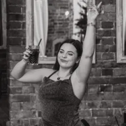 Kobieta z drinkiem w błyszczącej sukience, pozująca na tle ceglanej ściany we Wrocławiu, z uniesioną ręką i uśmiechem.