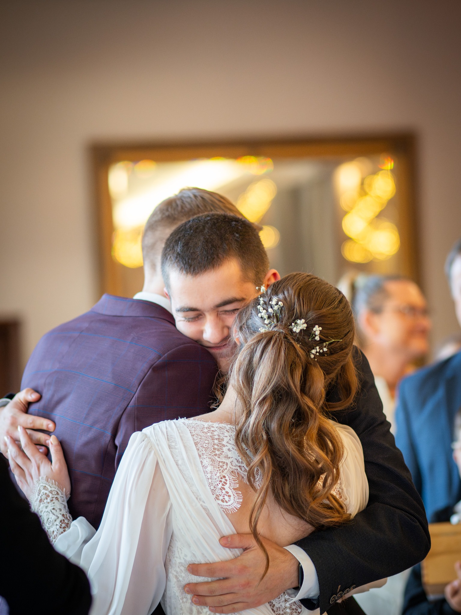 Uścisk nowożeńców z gościem weselnym, panna młoda w sukni z koronkowymi detalami, pan młody w bordowym garniturze w kratę, w tle rozmyte twarze gości i lustro.